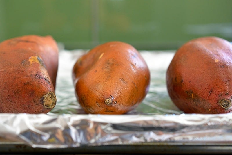 Three raw sweet potatoes on a aluminum foil-lined rimmed baking sheet.