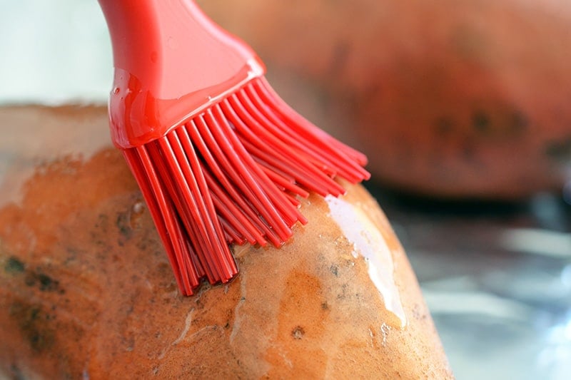 Close-up of a silicone brush applying melted fat on top of a raw sweet potato.