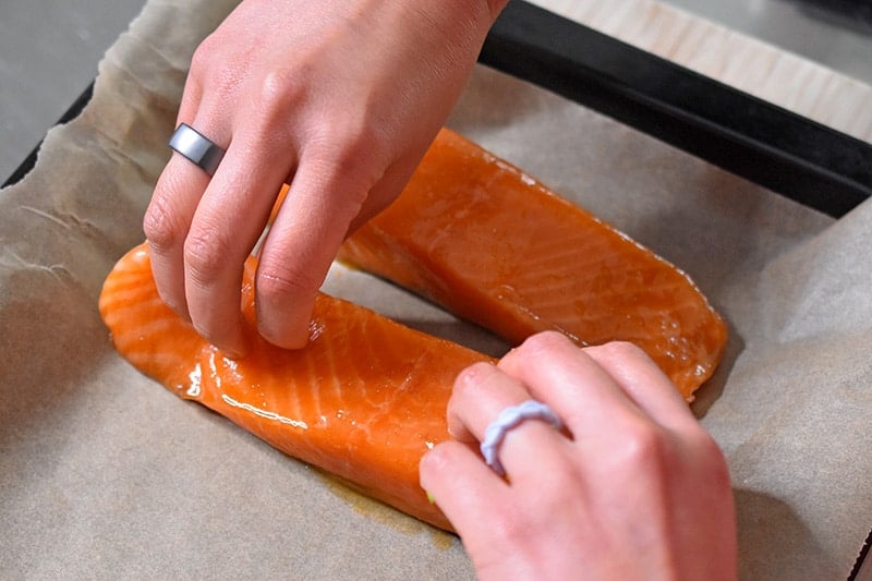Two hands are placing the marinated teriyaki salmon skin-side down on a parchment paper lined baking sheet.