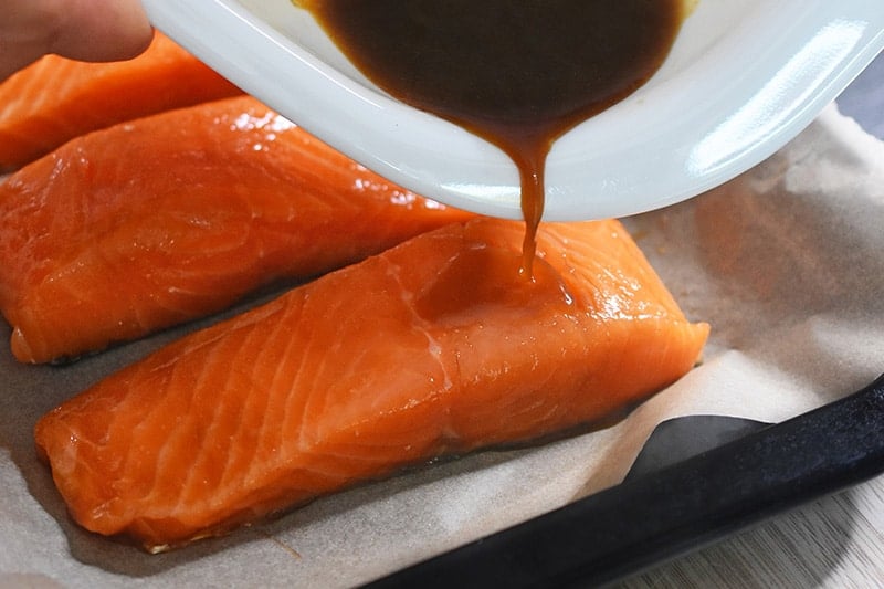 Someone is pouring the marinade over the salmon fillets on the baking sheet.