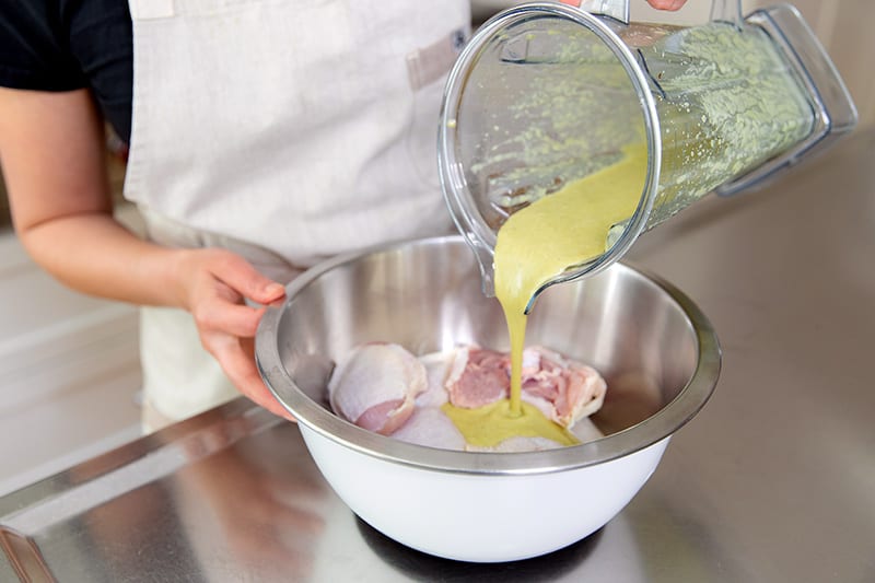 Pouring the paleo & Whole30 Asian chicken marinade over chicken thighs in a white stainless steel bowl.