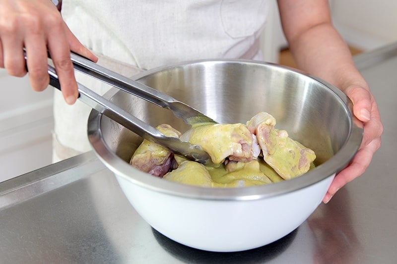 Tossing chicken thighs in a bowl filled with marinade with a pair of tongs.
