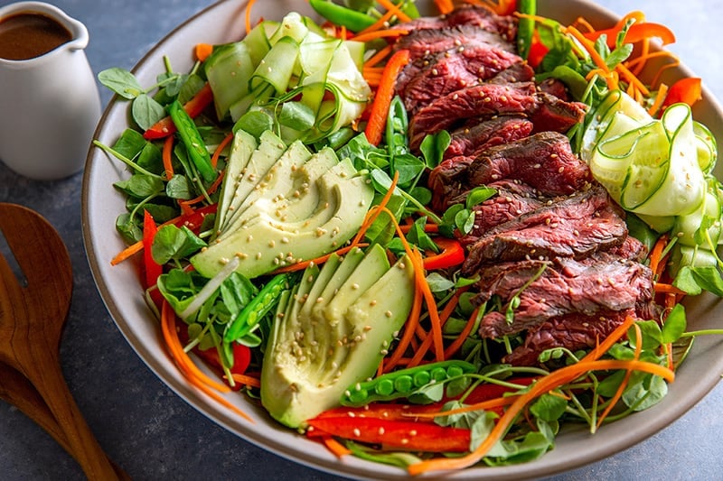 A side shot of a beautiful Whole30 Asian Steak Salad with a container of salad dressing on the side.