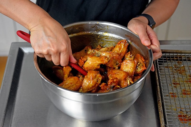 Someone tossing crispy oven chicken wings in sticky sauce with a maroon spatula.