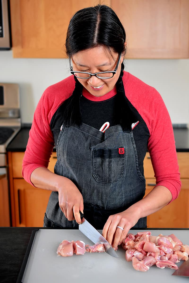 An Asian woman in a gray apron and red and black shirt is cutting boneless, skinless chicken thighs into uniform pieces. 
