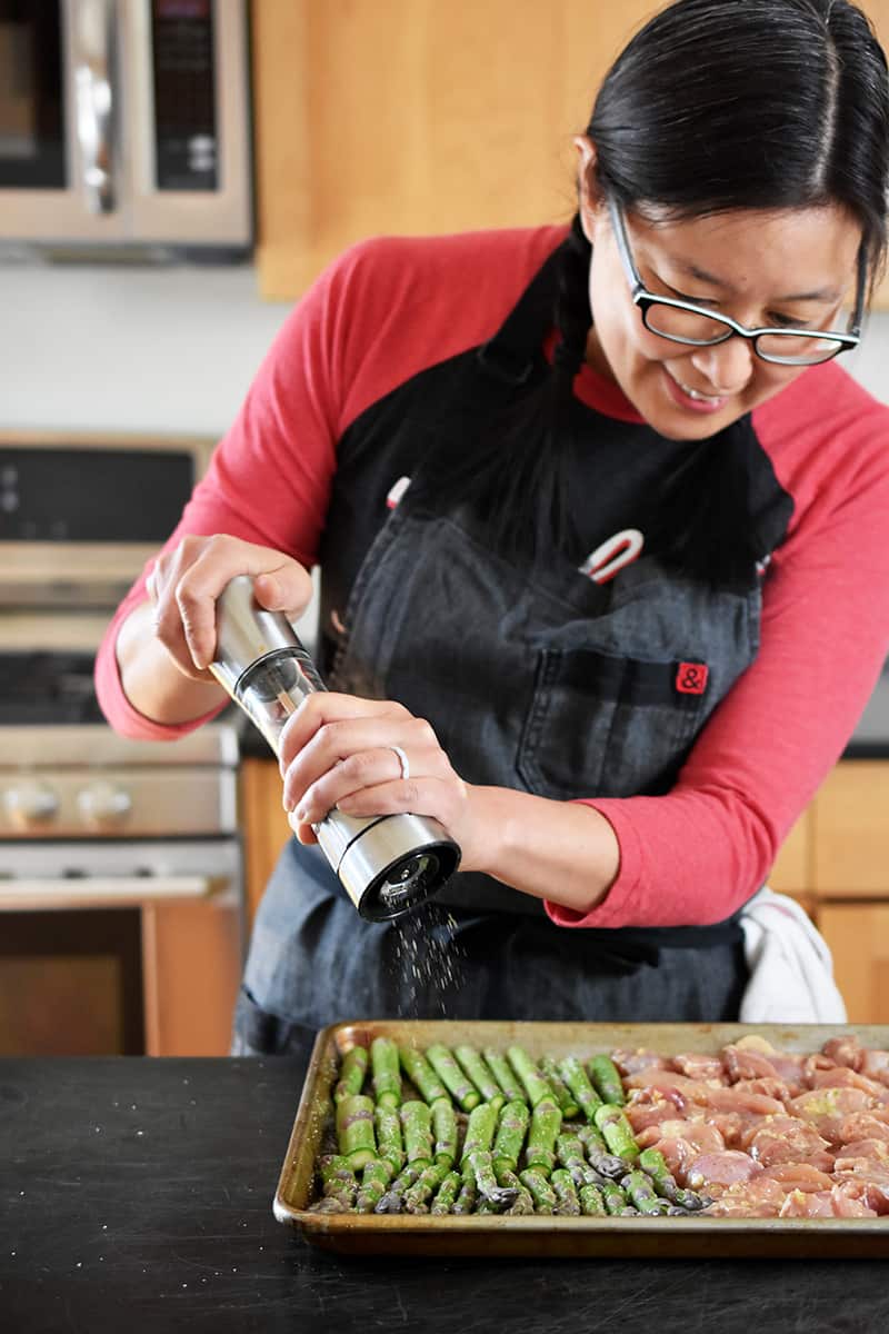 An Asian woman is adding freshly ground black pepper from a pepper mill onto some asparagus on a sheet pan.