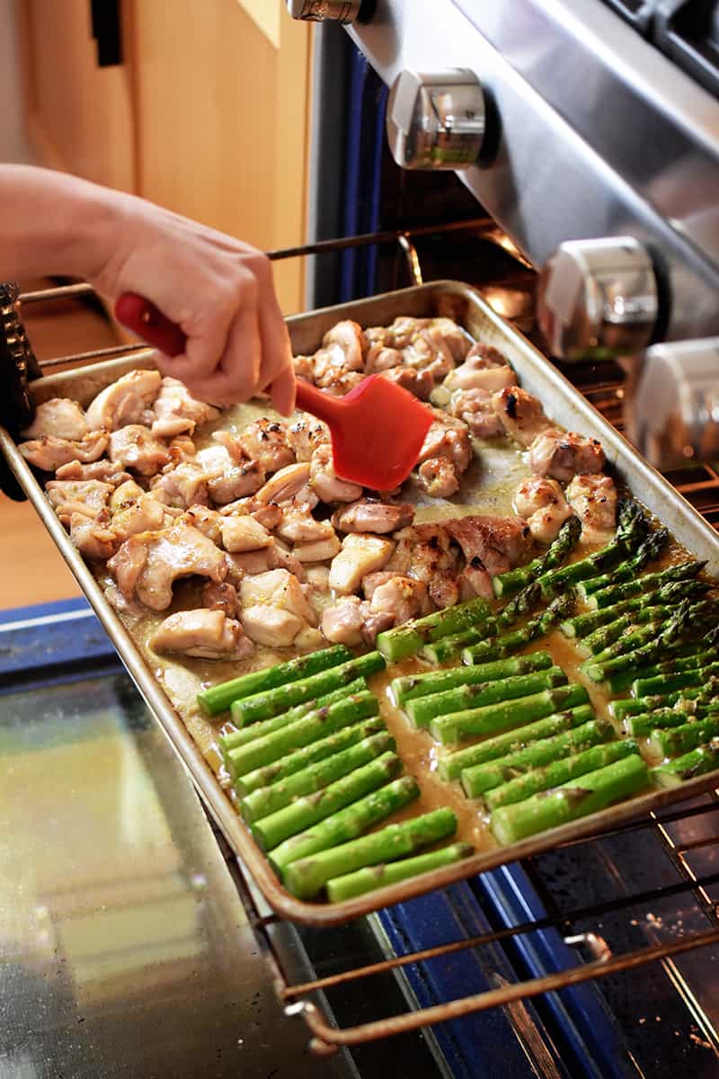 An open oven where someone is using a red silicone spatula to stir sheet pan chicken and asparagus at the halfway point.