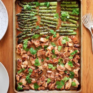 An overhead shot of sheet pan chicken and asparagus on a wooden table, next to bowls of cauliflower rice, plates, and forks.