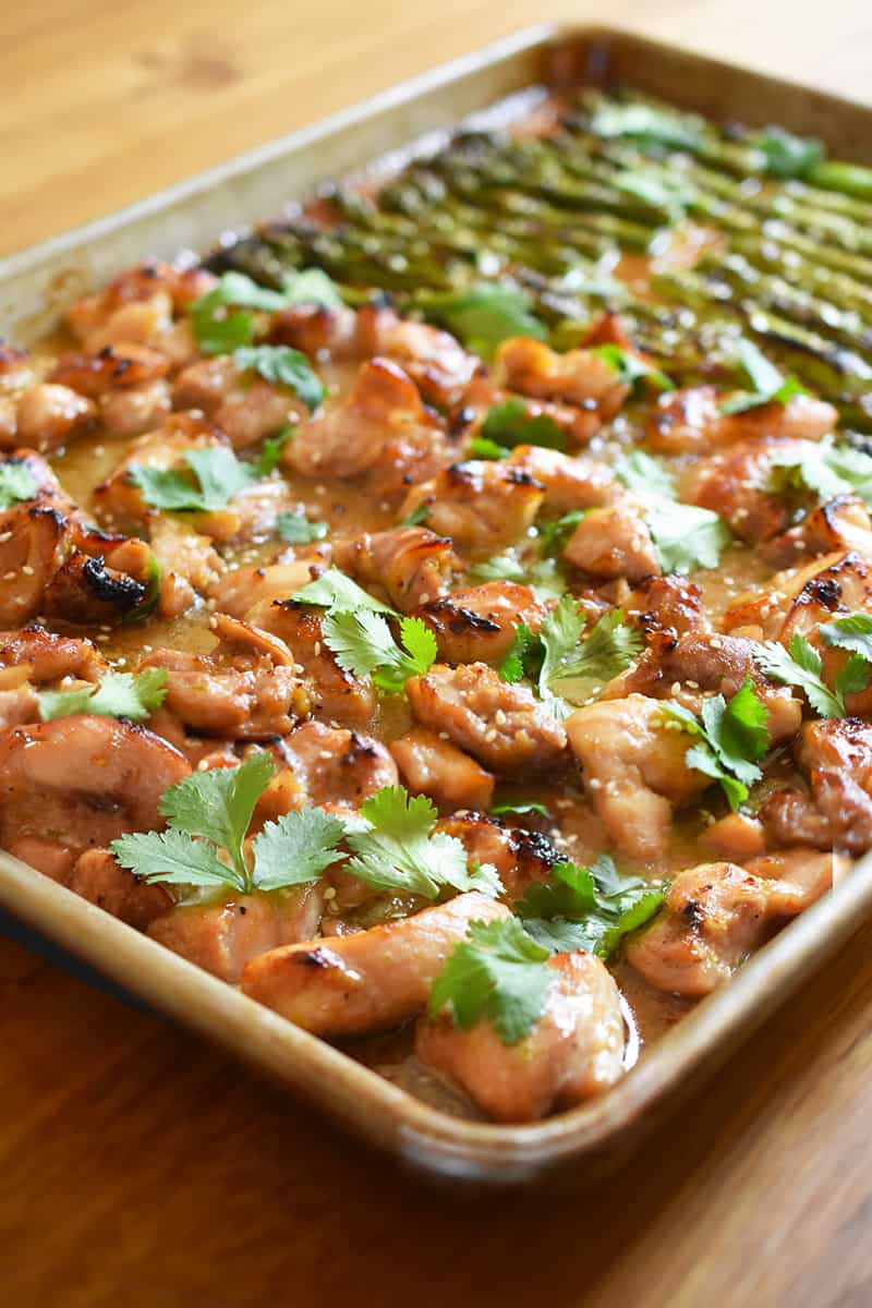 A side view of sheet pan chicken and asparagus right topped with cilantro and sesame seeds on a wooden tabletop.