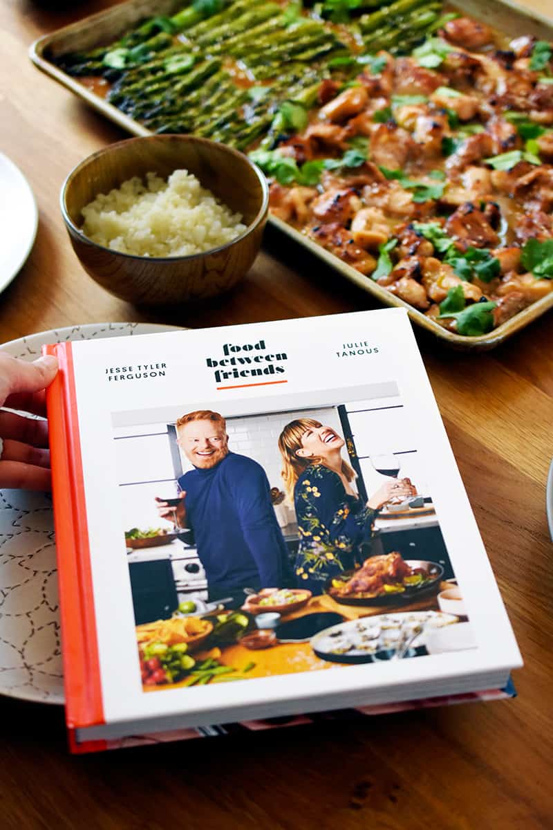 A hardcover copy of Food Between Friends cookbook is next to a Sheet Pan Chicken and Asparagus and a bowl of cauliflower rice.