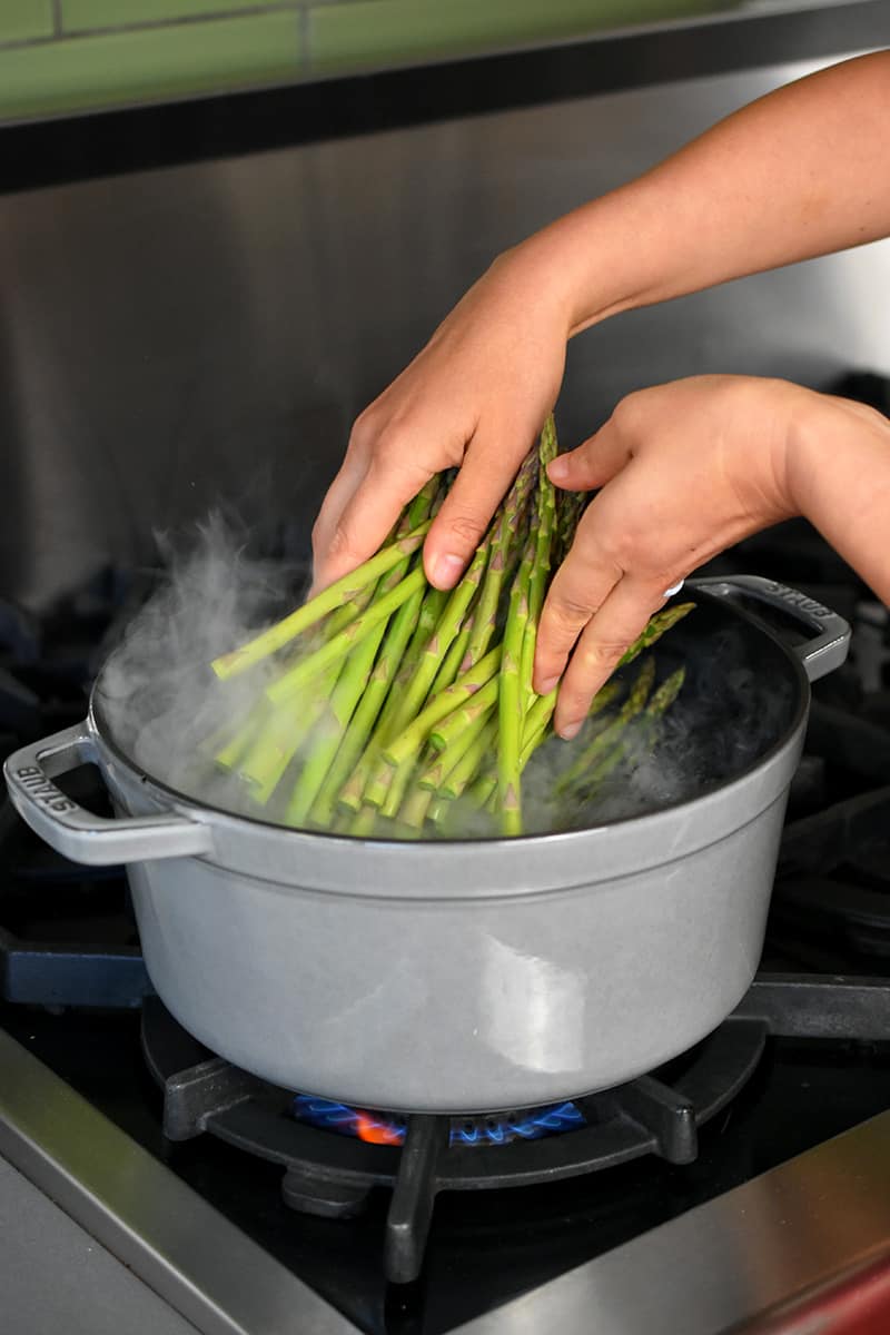 Two hands adding asparagus to a gray stock pot filled with boiling water.