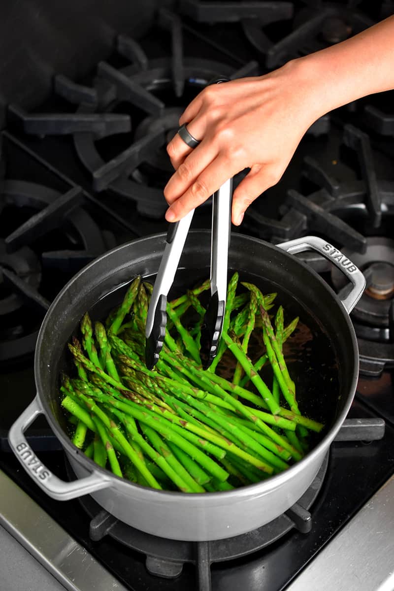 An overhead shot of a gray pot filled with boiling water and bright green asparagus. A hand with a pair of tongs is mixing them around.