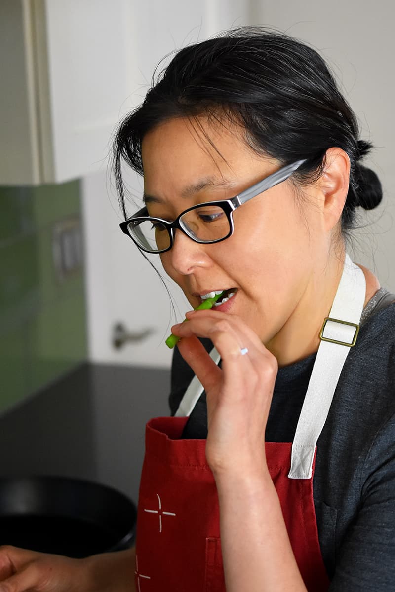 An Asian woman in a red apron is biting on a piece of asparagus.