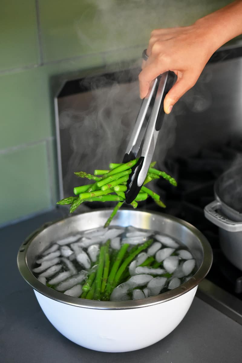 Someone using tongs to place cooked asparagus into a large bowl of ice water.