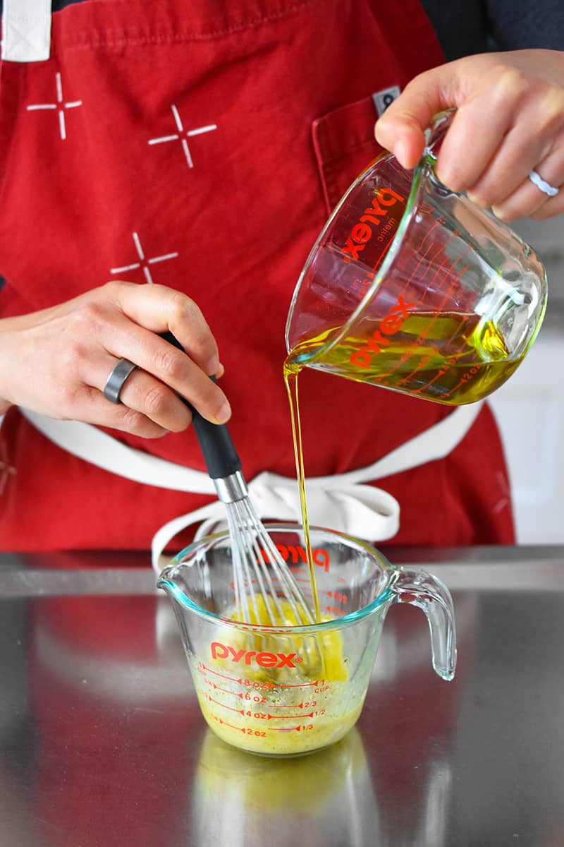 Someone in a red apron is whisking extra virgin olive oil into a measuring cup with vinaigrette ingredients.