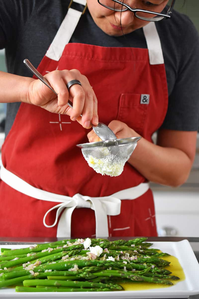A woman in a red apron is pressing egg whites through a small sieve on top of cooked asparagus