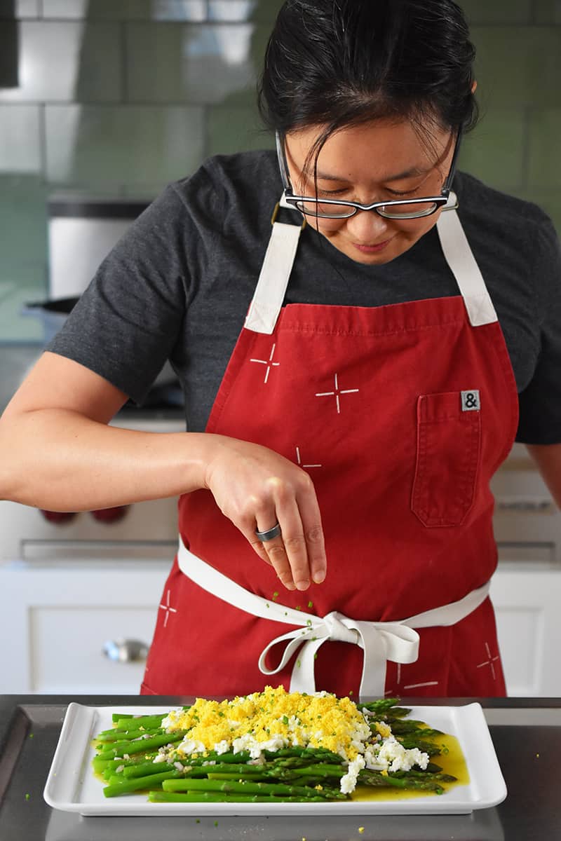 An Asian woman in a red apron is sprinkling chopped chives on top of asparagus mimosa.