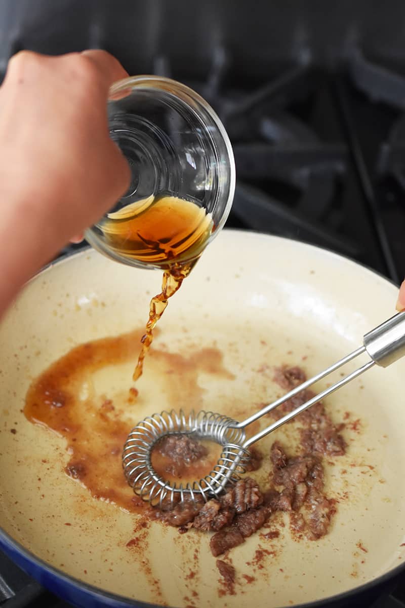 A hand is adding a bowl of sherry vinegar into a ceramic cast iron skillet filled with minced anchovies and garlic