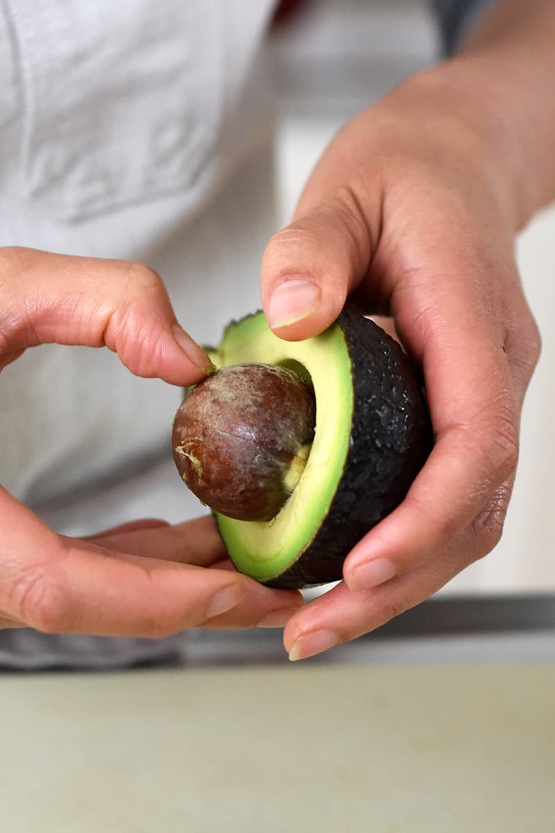 A person in a white apron is pulling an avocado pit from a halved avocado