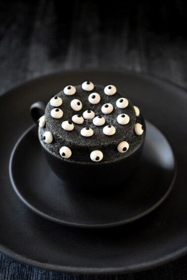 A black mug on a two black plates is filled with a black sesame cake topped with candy eyeballs.