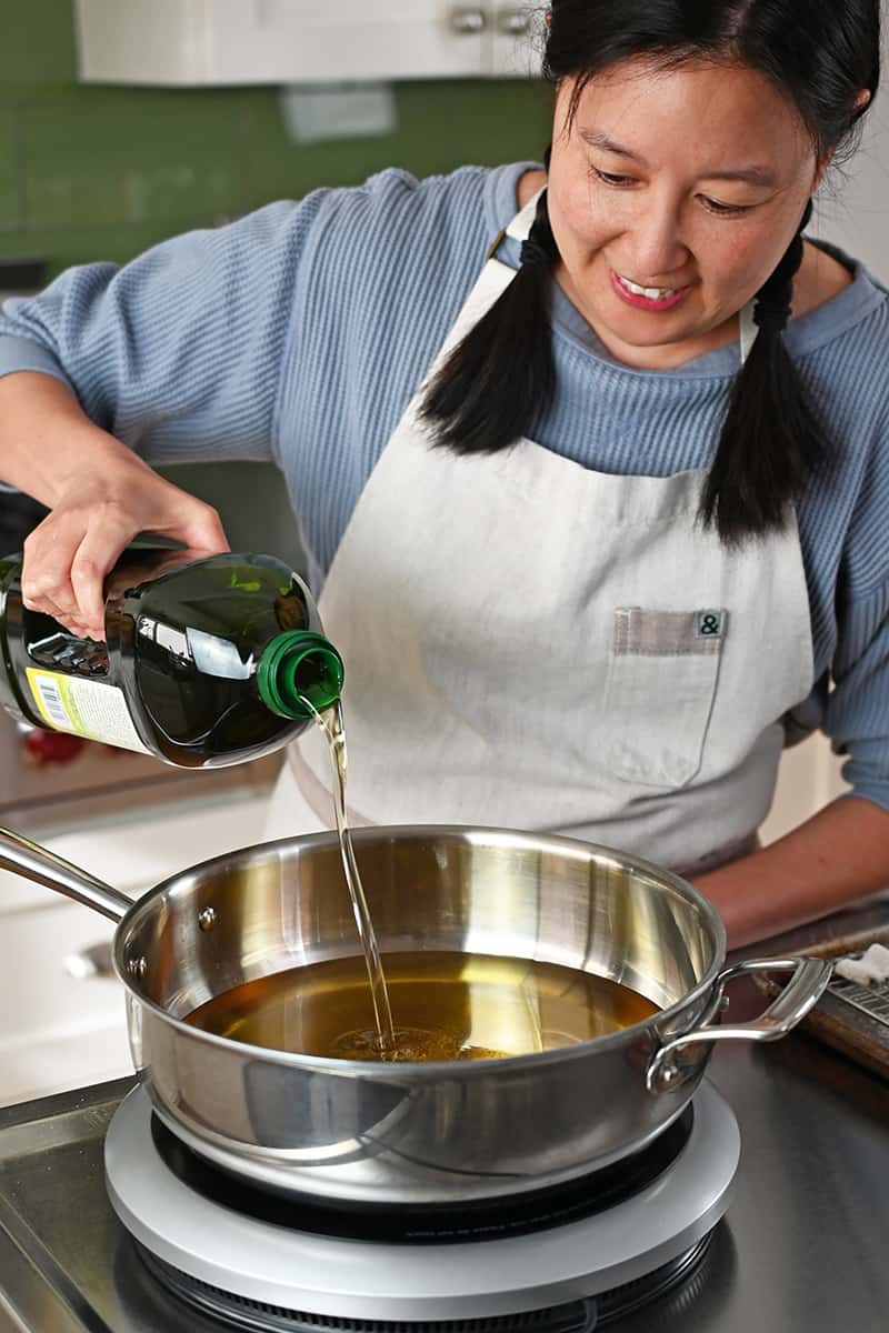 A smiling Asian woman is pouring avocado oil into a pot on an induction cooktop.
