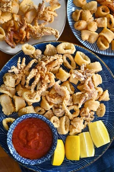 An overhead shot of three plates with paleo and gluten-free fried calamari. There is a small bowl with marinara sauce in it and lemon wedges on the side