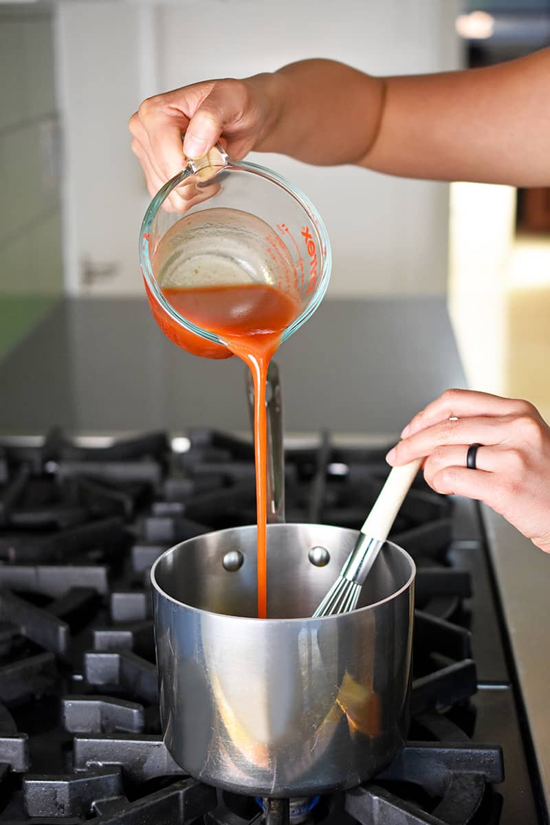 A hand is pouring cayenne pepper sauce from a liquid measuring cup into a small saucepan to make the buffalo cauliflower sauce