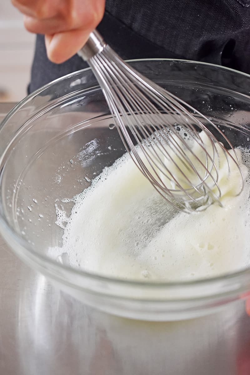 A whisk is whipping egg whites until frothy and bubbly in a clear glass bowl.