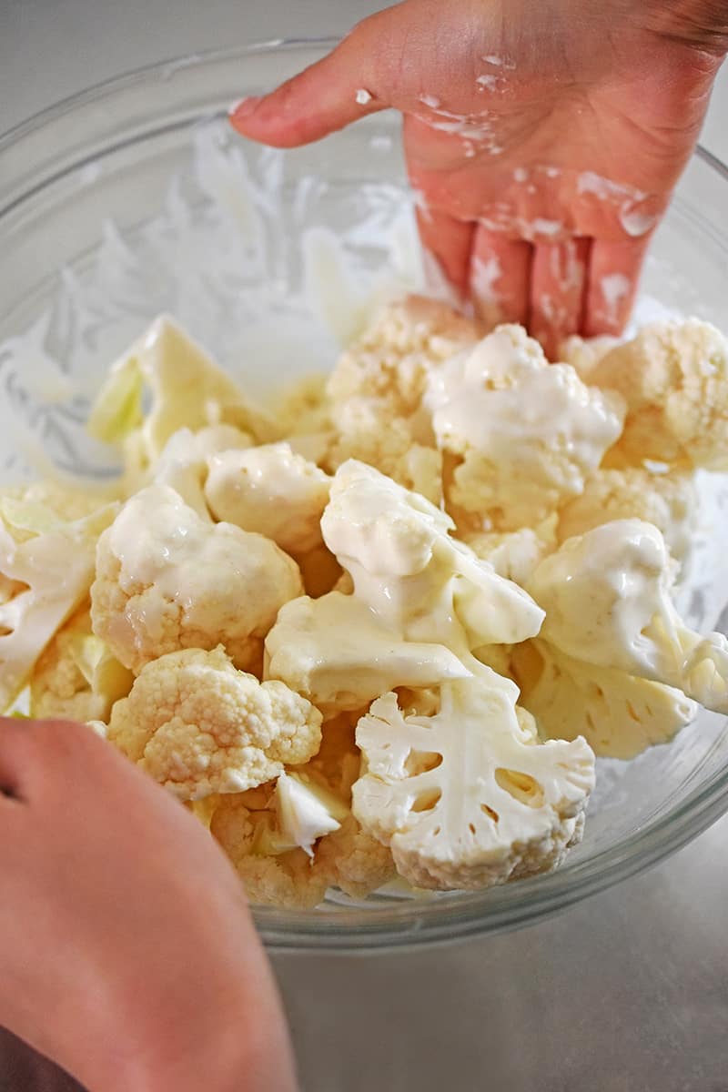 Two hands are tossing cauliflower florets in the gluten-free, paleo, and Whole30 batter for baked buffalo cauliflower.