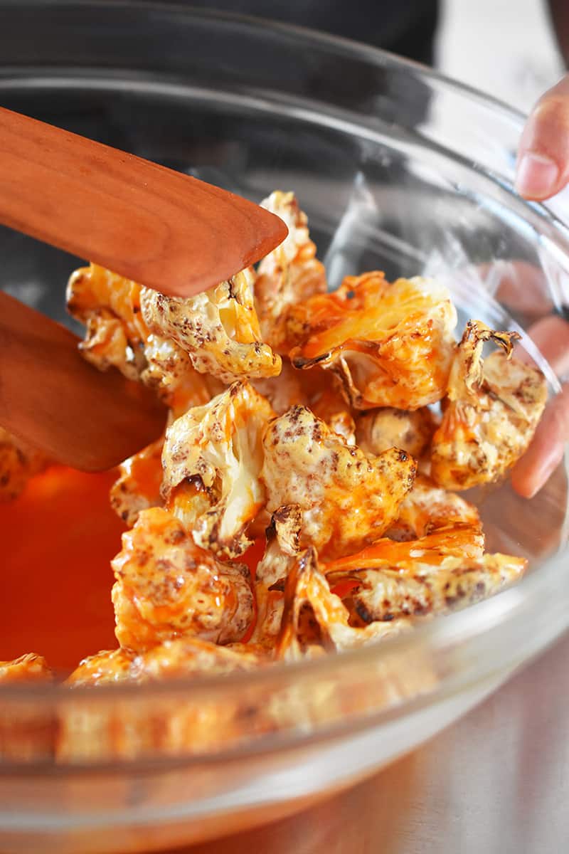 Tossing crispy cauliflower florets with buffalo sauce in a clear glass bowl.