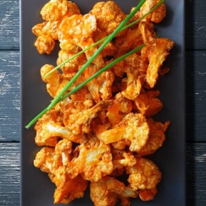 An overhead shot of baked buffalo cauliflower on a rectangular gray plate. There are three chives on top.