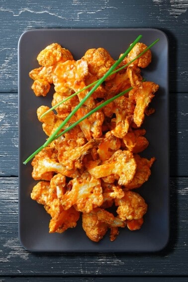 An overhead shot of baked buffalo cauliflower on a rectangular gray plate. There are three chives on top.