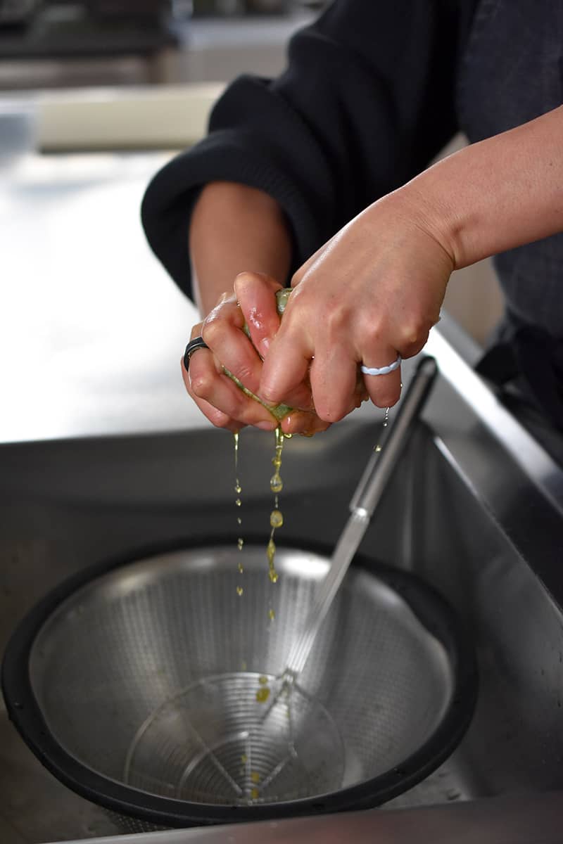 Two hands are wringing the liquid out of the cooked kale wrapped in a piece of cheesecloth.