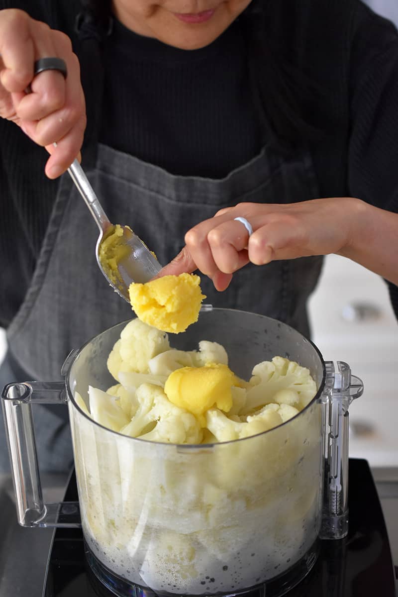 Adding ghee to an open food processor with cooked cauliflower florets.