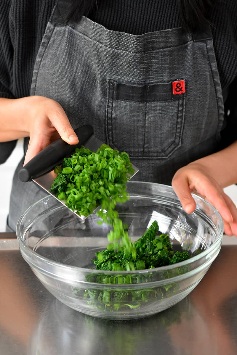 A person in a gray apron is transferring chopped kale and scallions with a bench scraper into a large glass bowl.