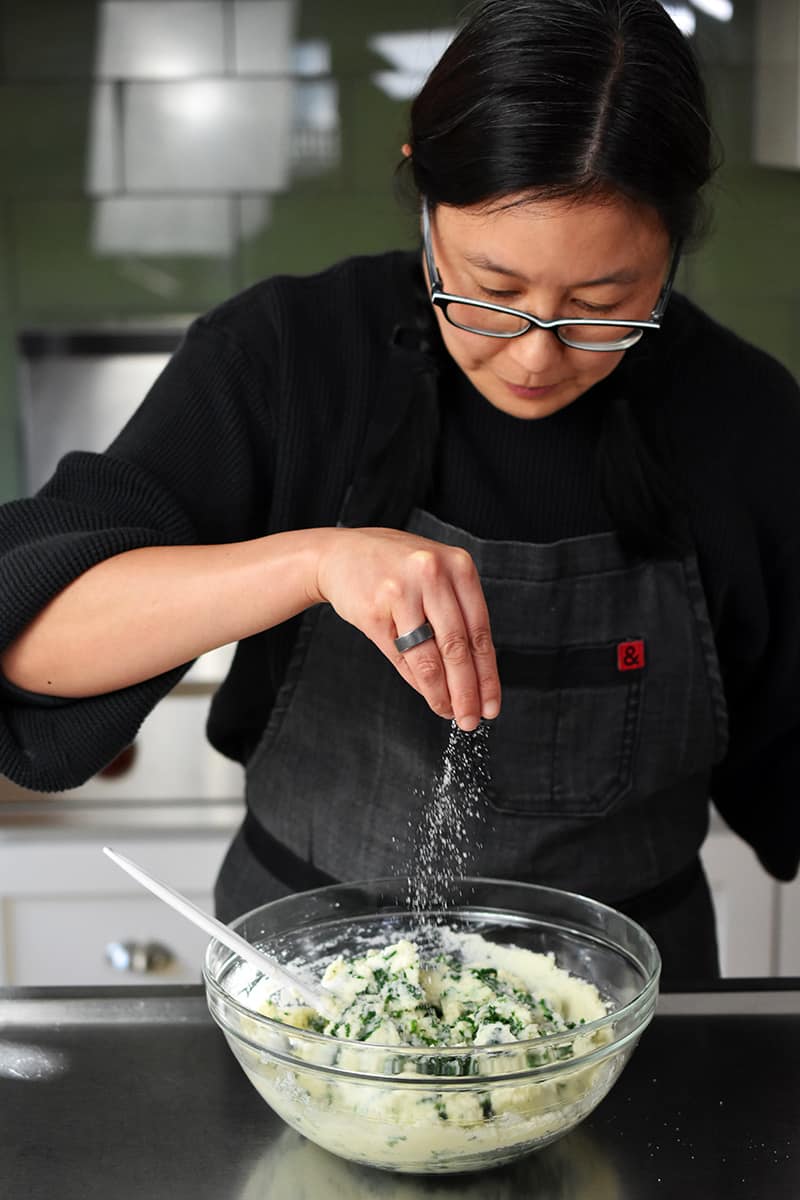 An Asian woman in glasses is sprinkling salt on some cauliflower colcannon in a bowl.
