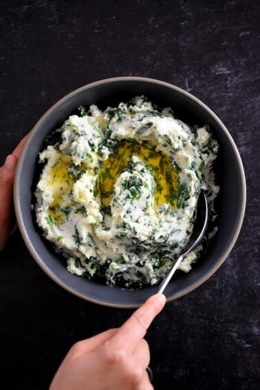 A hand is scooping paleo cauliflower colcannon from a gray bowl. The colcannon is white pureed cauliflower with flecks of green kale and scallions in it. There is melted ghee on top.