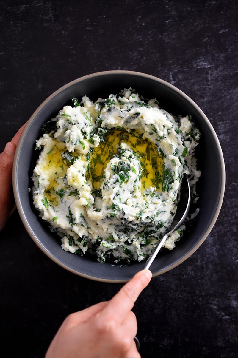 A hand is scooping paleo cauliflower colcannon from a gray bowl. The colcannon is white pureed cauliflower with flecks of green kale and scallions in it. There is melted ghee on top.