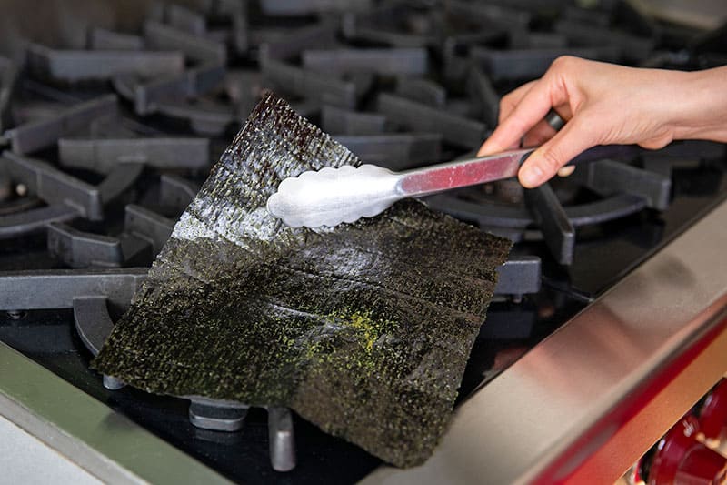 Using a pair of metal tongs to fan a piece of nori over a gas burner to toast it for California sushi hand rolls.