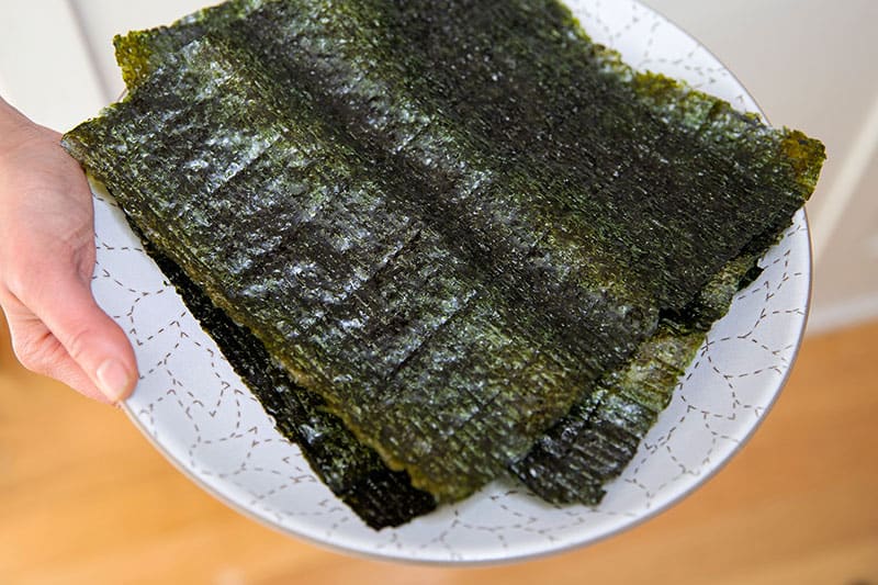 Someone holding a white plate with toasted nori sheets cut in half to make California hand rolls or Temaki.