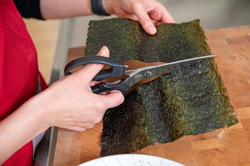Someone using kitchen shears to cut a toasted sheet of nori in half width-wise to make Whole30 and keto California hand rolls.