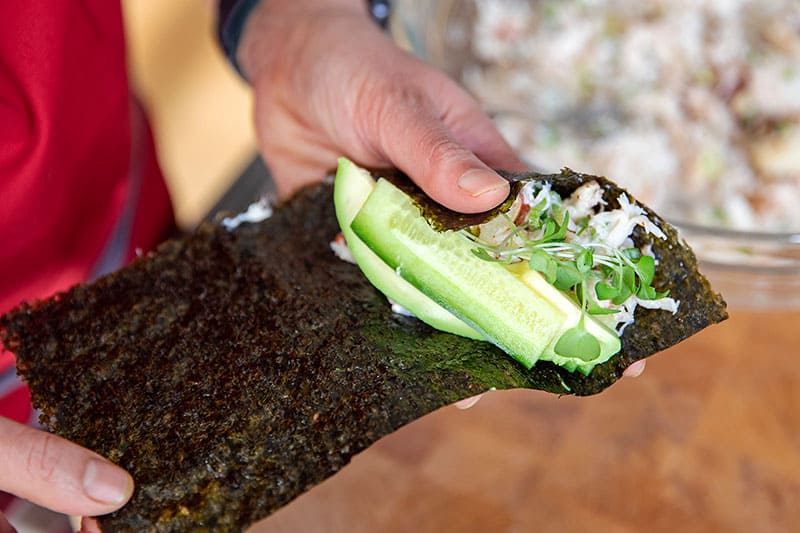 Adding the sliced cucumber, avocado, and sprouts on top of the crab salad on a piece of nori.
