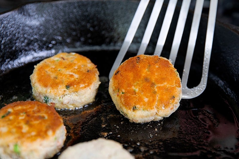 Flipping golden brown paleo crab cakes in a cast iron skillet with a metal fish spatula.