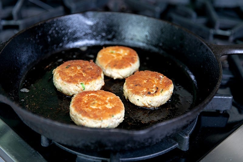 Four golden brown gluten-free crab cakes frying in a cast iron skillet.
