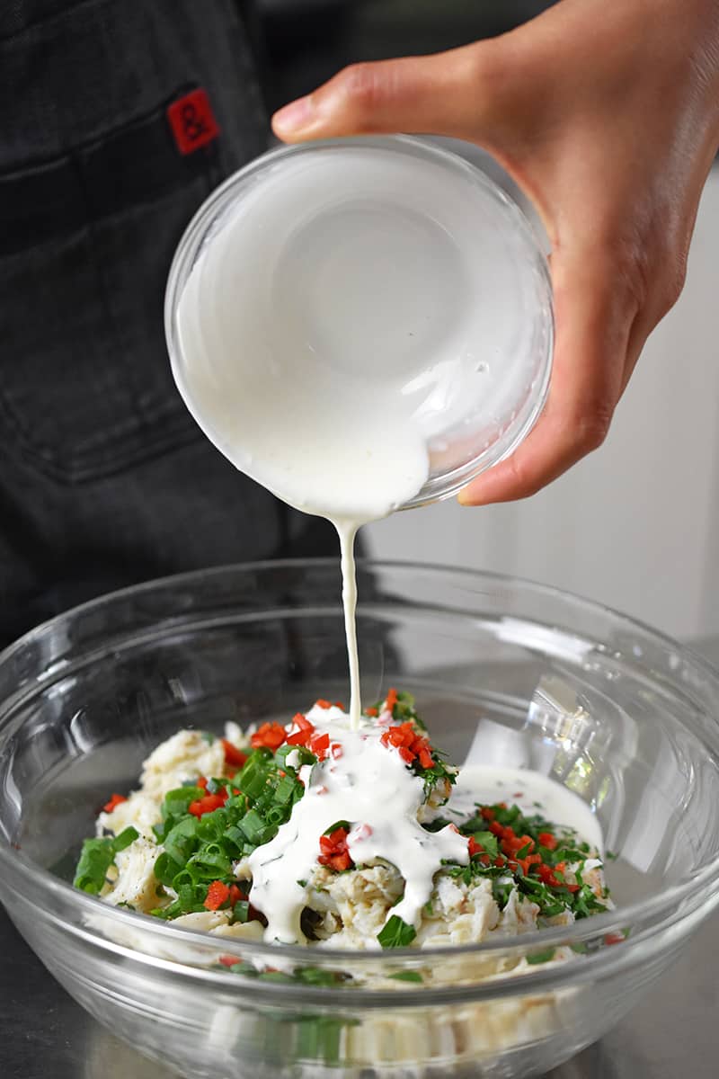 Pouring the dressing on a bowl filled with crab salad ingredients.