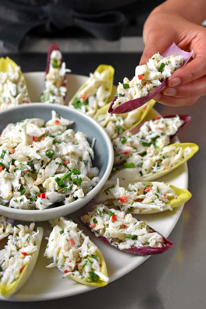 A hand is grabbing an endive spear filled with crab salad from a serving platter.