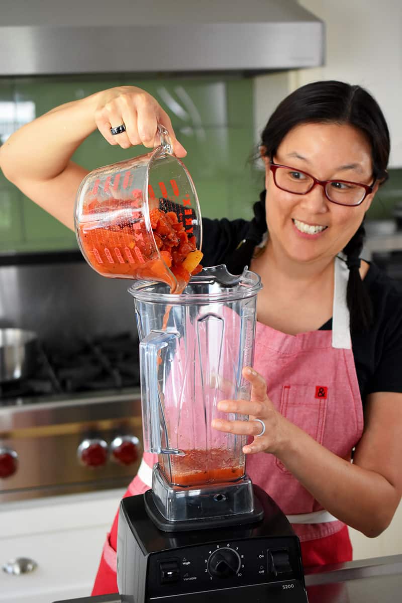 An Asian woman is pouring diced canned tomatoes into a Vitamix blender
