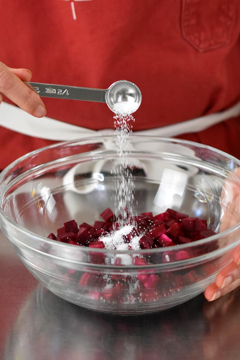 Someone in a red apron is adding a spoonful of salt to a clear bowl filled with diced purple beets
