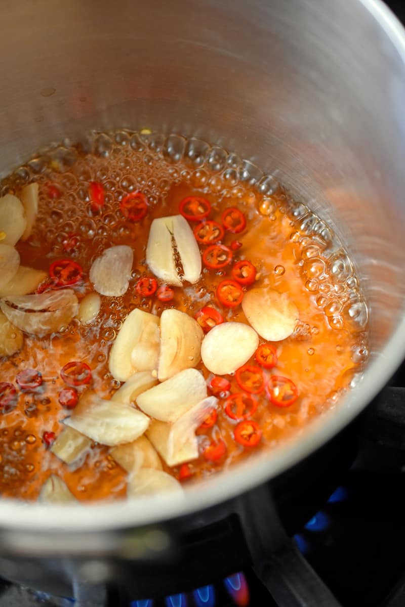 An overhead shot of a small saucepan filled with simmering vinegar, Thai chili peppers, and sliced garlic.