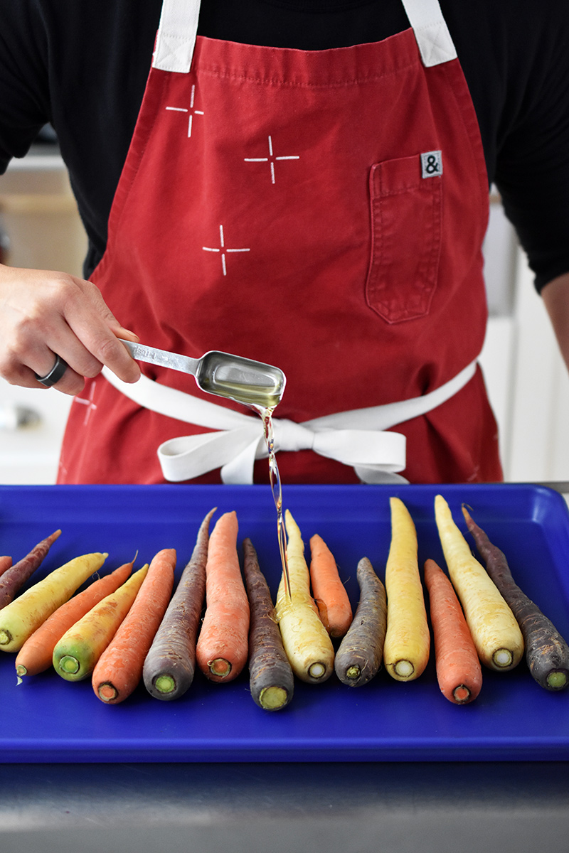 A person in a red apron is drizzling avocado oil on a rimmed baking sheet filled with colorful carrots.